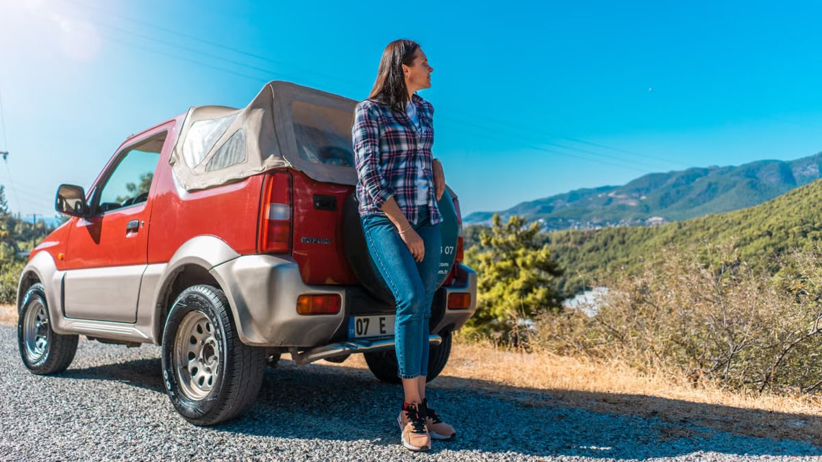 A woman leans against a red car on a mountain road in Alanya, Turkey, enjoying a sunny day.